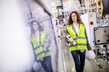 Mitarbeiterin in Warnweste und mit Helm und Aktenordner in der Hand geht durch eine Produktionshalle