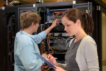 Zwei Studierende stehen an Servertower, der Junge steckt Kabel ein, das Mädchen notiert auf Klemmbrett