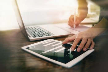 Person sitzt an einem Laptop und arbeitet gleichzeitig an daneben liegendem Tablet